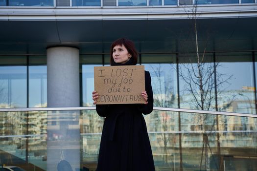 Woman holds sign saying I lost my job because of coronavirus. Concept of job loss due to the COVID-19 virus pandemic. Female with closed eyes stands against background of business cente