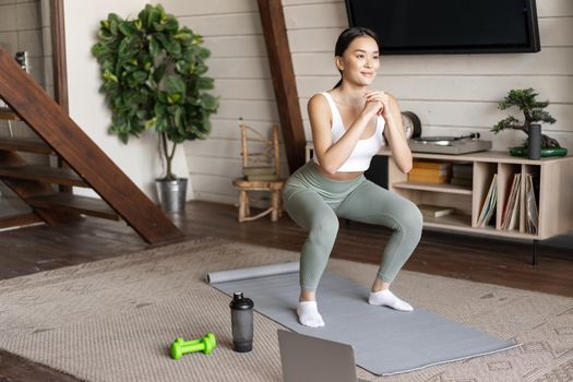 Young asian fitness girl doing squats workout at home, follow online sport gym instructor, standing on floor mat and smiling, exercising in living room.