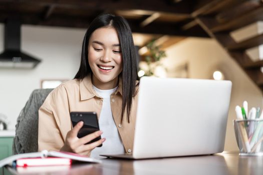 Smiling asian girl student doing homework on laptop. Woman using cellphone and computer for work from home.