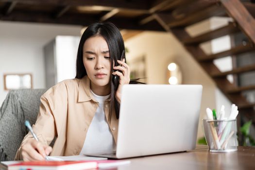 Image of upset asian woman talking on mobile phone and writing down notes on notebook with concerned face. Busy girl working from home, sits near laptop.