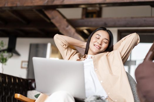 Smiling korean woman resting with hands behind her head, using laptop and relaxing at home.