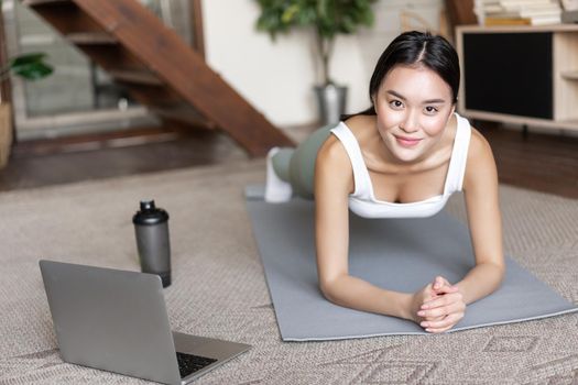 Sporty asian girl in activewear doing workout at home, follow video sport guide, online fitness instructor on laptop, standing in plank on floor mat.