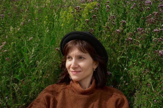Horizontal portrait of brunette smiling and looking away young adult lady wearing hat and sweater sitting in wildflower field Karelia