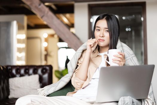 Sick woman sitting at home, watching videos on laptop and drinking hot tea.