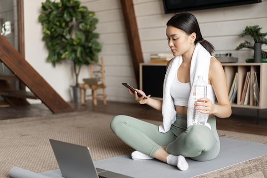 Asian girl in fitness clothing sitting at home on floor mat, resting after workout, checking mobile phone sport application.