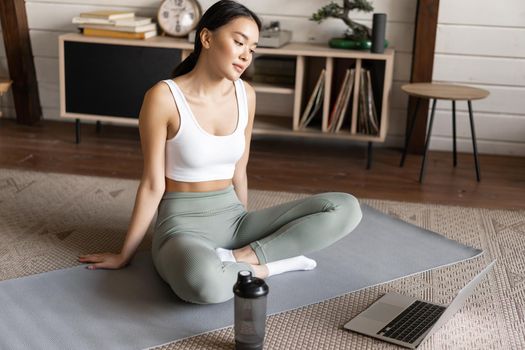 Sport and lifestyle. Young asian girl follow online trainer instructions, workout using video course on laptop, sitting on floor mat for exercises at home.