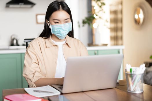 Asian girl in medical mask working from home while being sick with covid, using laptop.