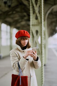 Railway station. Beautiful girl is waiting for train and looks at cell phone. Woman travels light. Middle-aged lady with dark hair in warm clothes in red hat and red bag at winter or autumn, Vertical