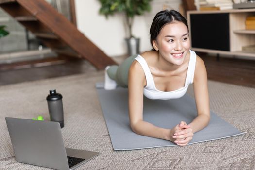 Sporty asian girl in activewear doing workout at home, follow video sport guide, online fitness instructor on laptop, standing in plank on floor mat.