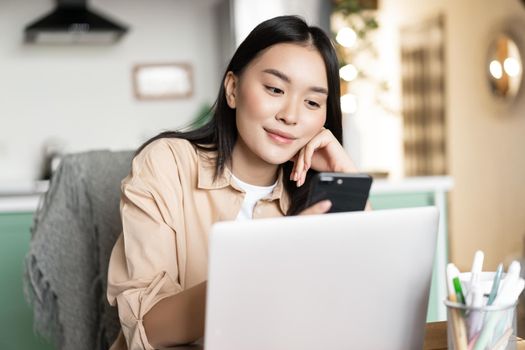 Smiling asian girl studying online, working on laptop and smartphone from home.