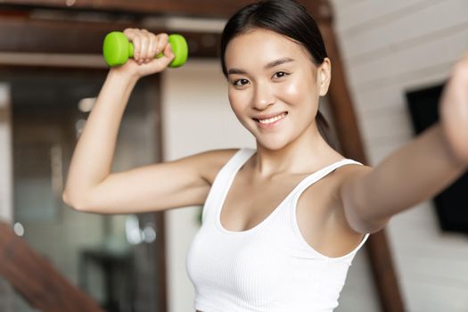 Smiling korean girl taking selfie with dumbbell, workout at home during pandemic, wearing activewear.