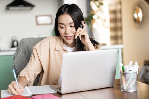 Asian woman working from home, answer phone call and writing down in notebook. Girl entrepreneur busy on cellphone, sitting near laptop.