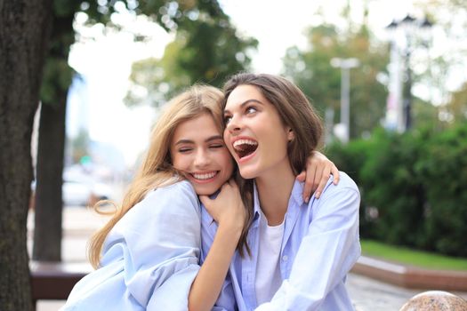 Two young smiling hipster women in summer clothes posing on street.Female showing positive face emotions