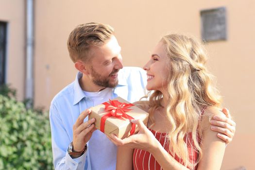 Astonished excited couple in summer clothes smiling and holding present box together while standing on city street.