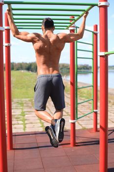 Muscular guy with naked torso pulling up on horizontal bar outdoor.