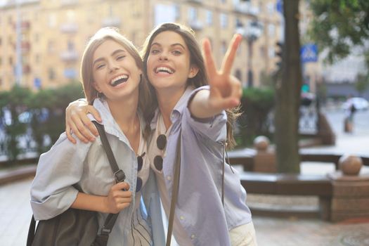 Two young smiling hipster women in summer clothes posing on street.Female showing positive face emotions