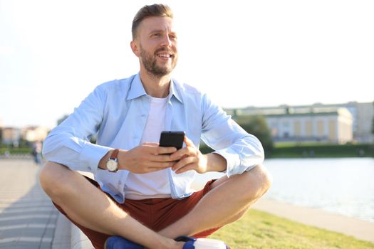 Happy young handsome man sitting on the bench outdoors and using smartphone.