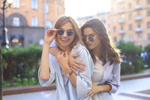 Two beautiful smiling girls in trendy summer clothes posing on street background. Models are having fun and hugging