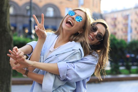 Two beautiful smiling girls in trendy summer clothes posing on street background. Models are having fun and hugging
