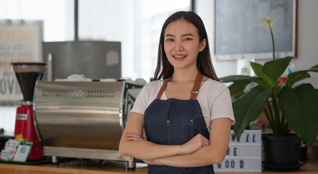 Beautiful asian woman store owner with standing in her coffee shop looking at camera and smiling.Portrait of girl waitress wearing apron and standing in front