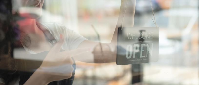 Portrait of smiling young barista girl in apron holding open sign board while standing at her cafe. elegant asian coffee shop female staff turn door plate in the morning in own store small business.