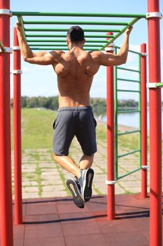 Muscular guy with naked torso pulling up on horizontal bar outdoor.