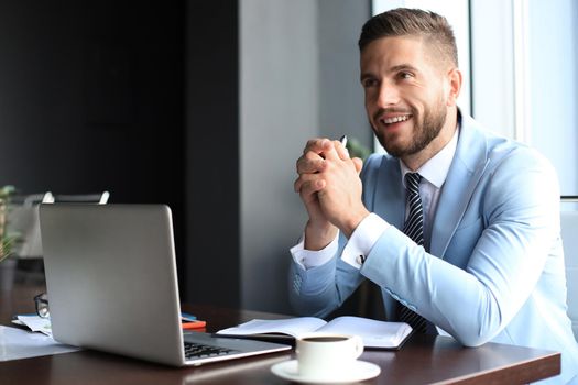 Portrait of happy businessman sitting at office desk, looking at camera, smiling.