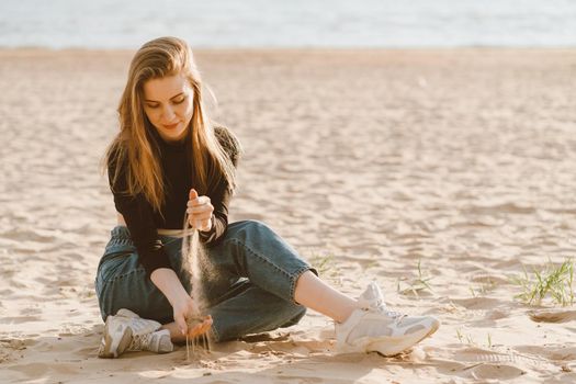 Full length beautiful woman sitting on beach at sunset in evening and pour sand. Slow life, social detox and self-immersion, single. Pretty female with long brown hair in casual clothes.