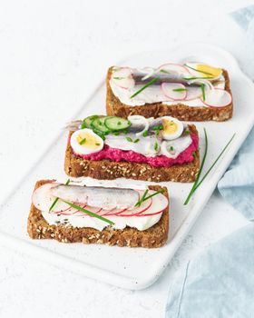 Savory smorrebrod, set of traditional Danish sandwiches. Black rye bread with anchovy, beetroot, radish, eggs, cream cheese on grey plate on a white stone table, side view, vertical