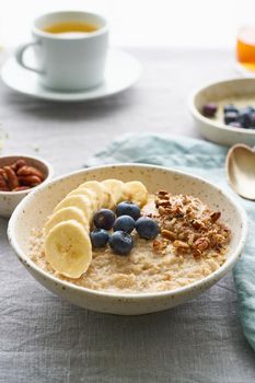 Whole oatmeal, large bowl of porridge with banana, blueberries, nuts for breakfast, morning meal. Side view, close up, vertical, gray table. Vegan tasty healthy diet.