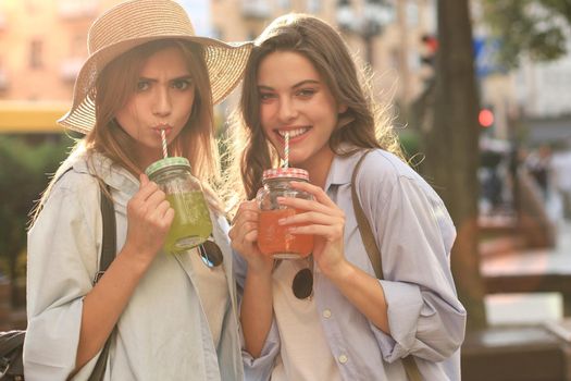 Two young beautiful smiling woman in summer clothes are having fun in city and drinking fresh cocktail