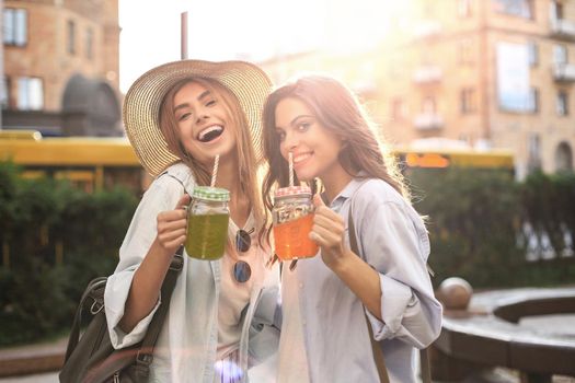 Two young beautiful smiling woman in summer clothes are having fun in city and drinking fresh cocktail