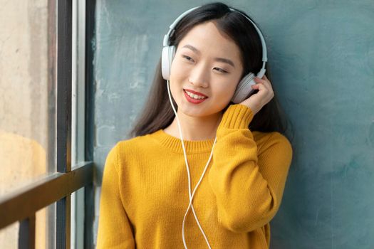 Young beautiful Asian girl listening to music with headphones sitting on window and looking out. Smiling pretty women