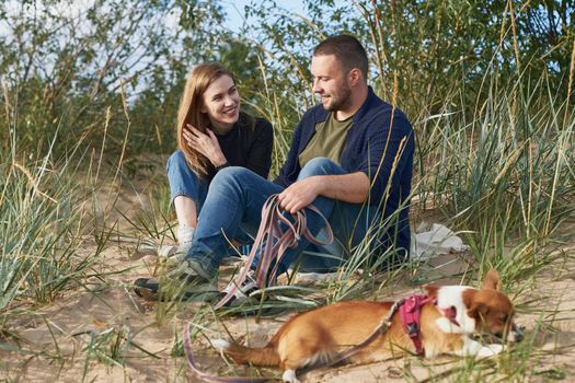 Young happy couple with corgi dog siting at sand. Handsome man and beautiful woman in casual clothes enjoying sunny afternoon on beach