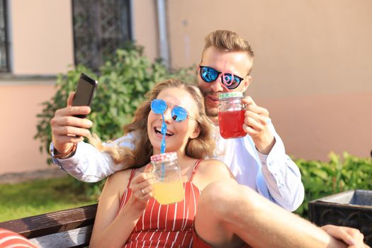 Romantic young couple in summer clothes smiling and taking selfie while sitting on bench in city street.