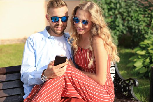 Romantic young couple in summer clothes smiling and using mobile while sitting on bench in city street.