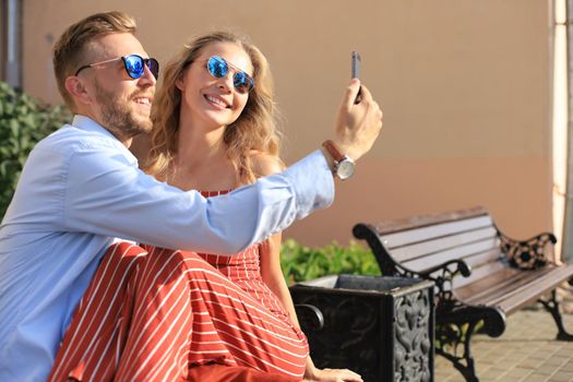 Romantic young couple in summer clothes smiling and taking selfie while sitting on bench in city street.