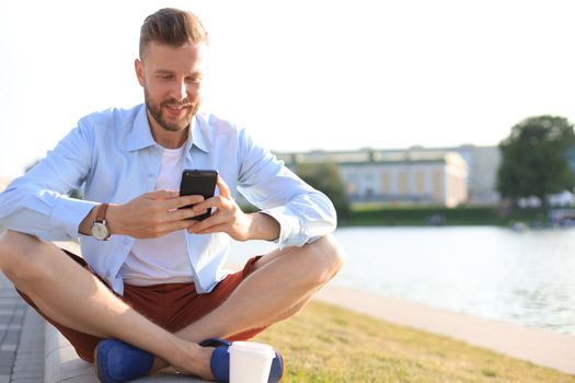 Happy young handsome man sitting on the bench outdoors and using smartphone.