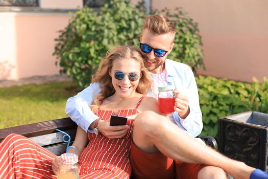 Romantic young couple in summer clothes smiling and taking selfie while sitting on bench in city street.