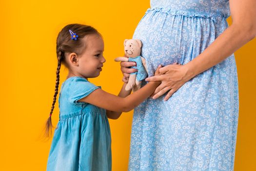 Motherhood, love, Childhood, pregnancy, hot summer - croped portrait pregnant unrecognizable mother woman blue dress little preschool Daughter girl sibling teddy bear hug mum belly on yellow background.