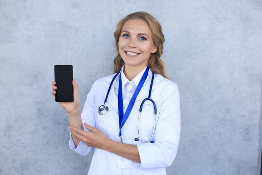 Female doctor smiling and showing a blank smartphone screen isolated over grey background