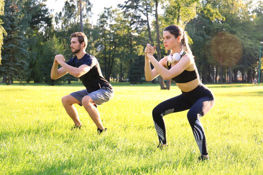 Young sporty man and woman doing workout and squatting together in green park during summer sunny day