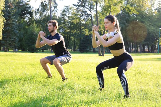 Young sporty man and woman doing workout and squatting together in green park during summer sunny day