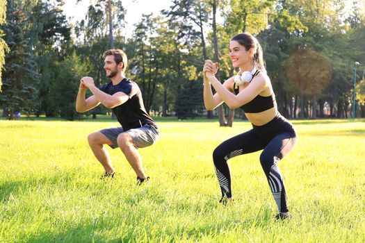 Young sporty man and woman doing workout and squatting together in green park during summer sunny day