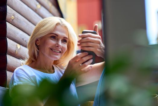 Pretty middle aged caucasian woman smiling to phone sitting on sunny terrace in apartment, selective focus. People spending time at home