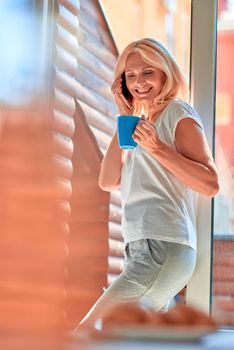 Smiling middle aged caucasian woman talking on phone while drinking coffee near sunny balcony in flat. People spending time at home