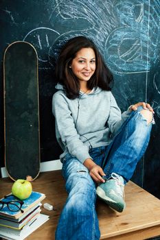 young cute teenage girl in classroom at blackboard seating on table smiling, modern hipster concept closeup