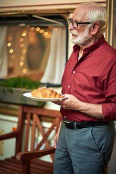 Side view of happy bearded elderly man standing near the camper van and holding a plate with croissant. Travel concept