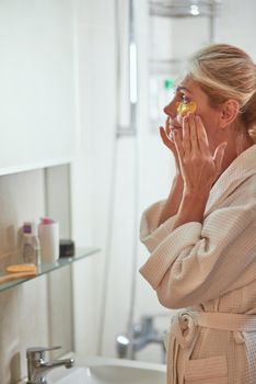 Middle aged caucasian woman in bathrobe using collagen anti-aging under eye patches standing sideways near mirror at home. Beauty, skincare and cosmetology concept