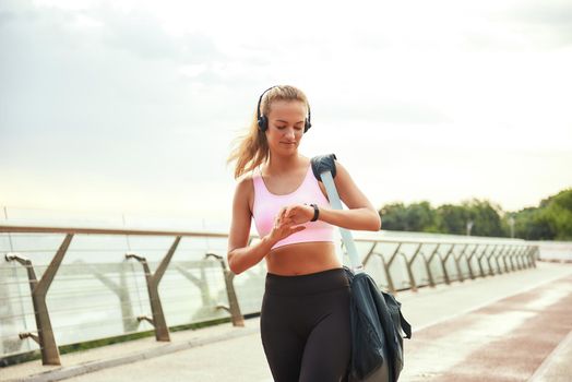 It was great workout Young sporty woman in headphones carrying bag and checking time on her watch while standing on the bridge. Motivation. Healthy lifestyle. Sport concept